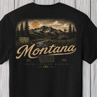 A black short-sleeve t-shirt with a vintage-style Montana graphic on the front, featuring a mountain landscape, evergreen trees, and the word 'Montana' in large script, displayed against a light gray wood plank background.