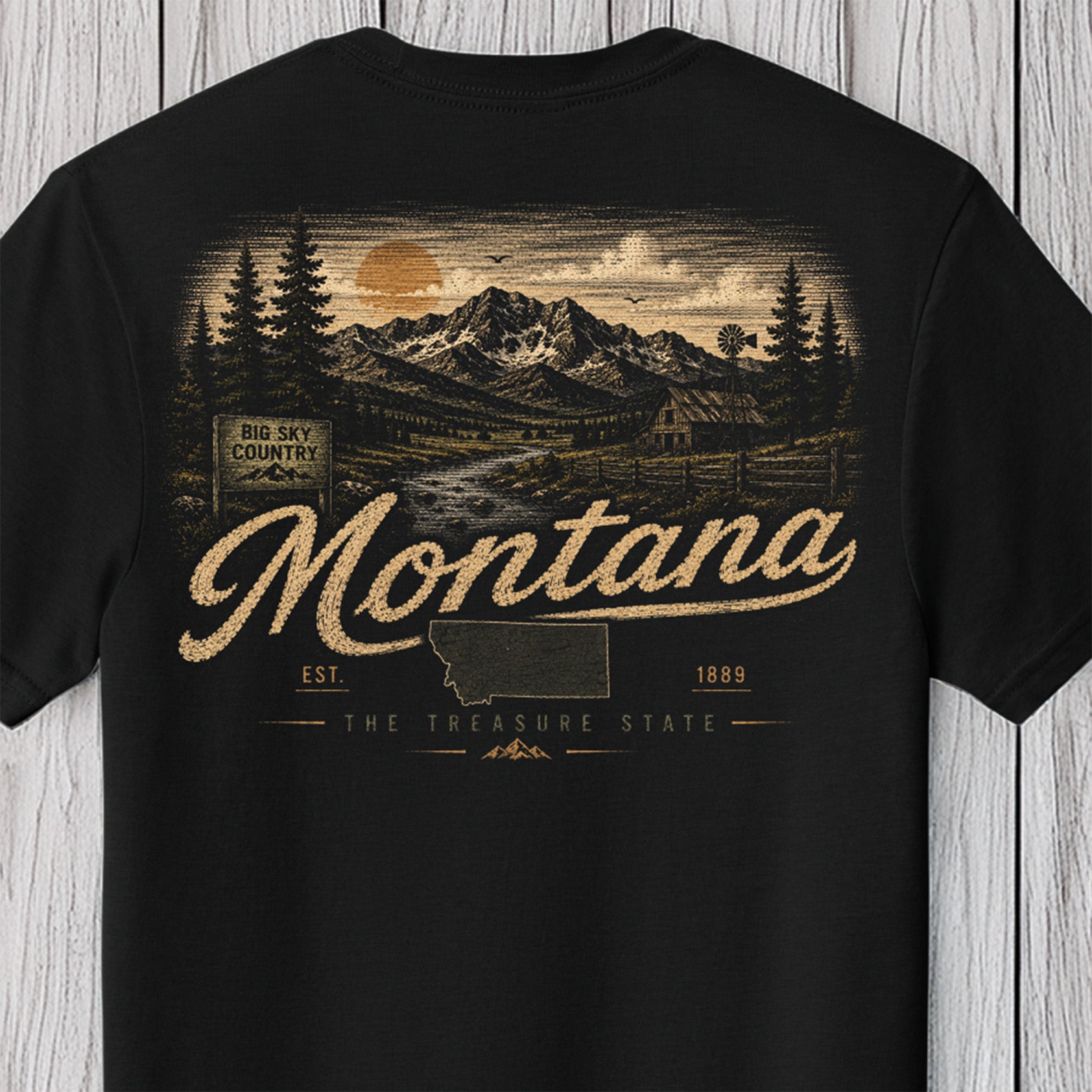 A black short-sleeve t-shirt with a vintage-style Montana graphic on the front, featuring a mountain landscape, evergreen trees, and the word 'Montana' in large script, displayed against a light gray wood plank background.