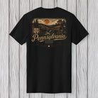 A black short-sleeve t-shirt shown from the back, featuring a vintage-style Pennsylvania graphic print in tan and orange tones, laid flat against a light gray wood plank background.