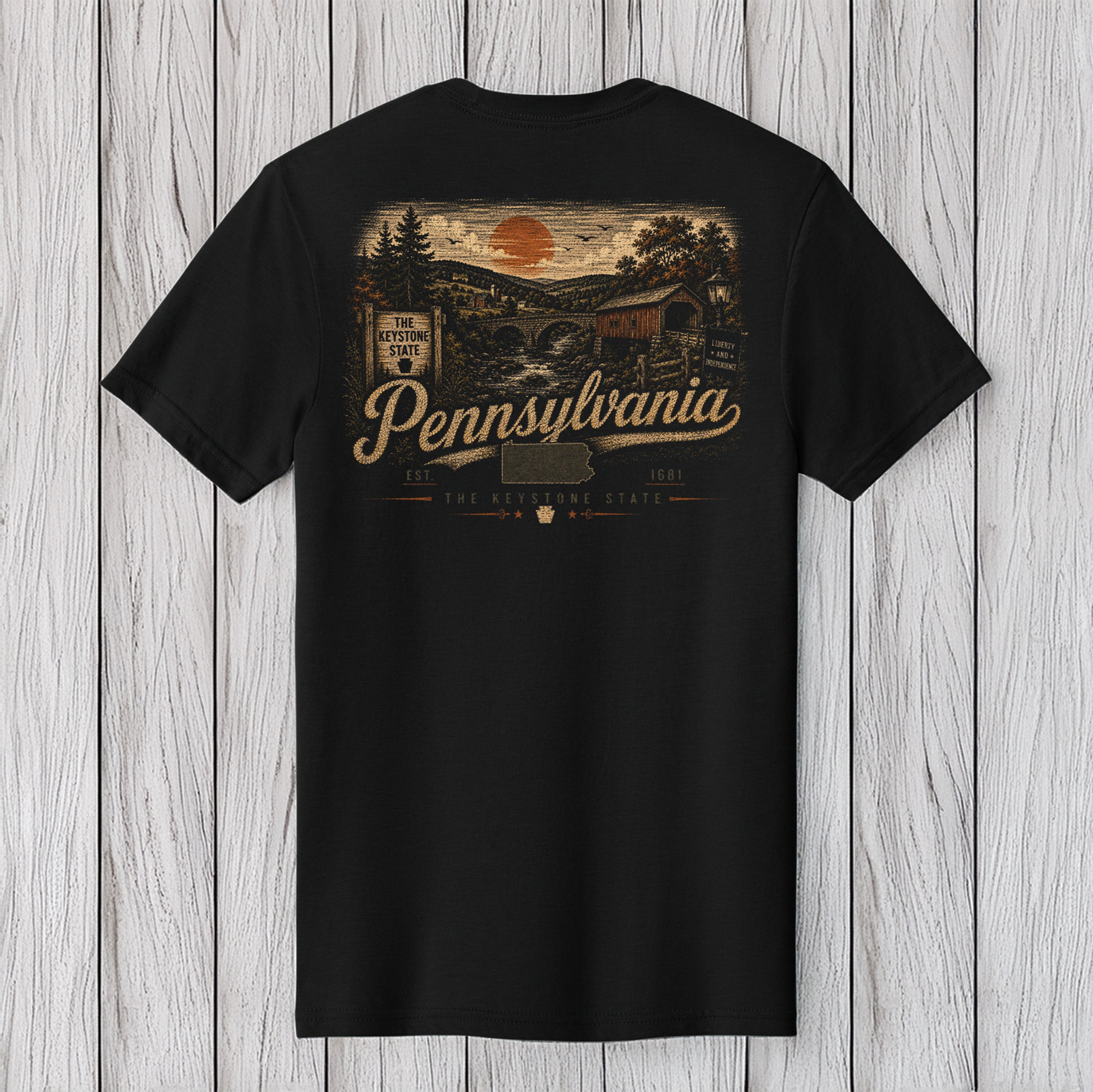 A black short-sleeve t-shirt shown from the back, featuring a vintage-style Pennsylvania graphic print in tan and orange tones, laid flat against a light gray wood plank background.