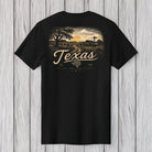 A black short-sleeve t-shirt with a vintage-style Texas landscape graphic on the front, featuring a sunset, trees, a barn, and the word 'Texas' in large script, displayed against a light gray wood plank background.