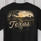 A black short-sleeve t-shirt featuring a vintage-style graphic print of a Texas landscape with a tree, barn, windmill, and the word 'Texas' in large script, displayed against a light wood plank background.
