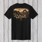 A black short-sleeve t-shirt with a vintage-style Utah graphic print on the front, displayed flat against a light gray wood plank background.