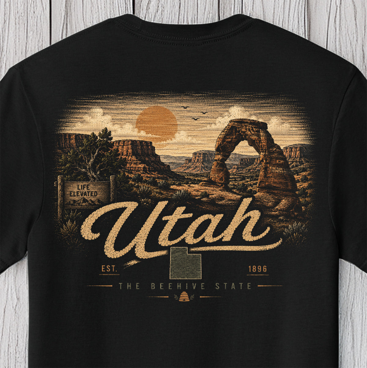A black short-sleeve t-shirt with a detailed graphic print of Delicate Arch National Park in Utah, featuring the text 'Utah' and 'The Beehive State' in a vintage style, displayed against a light gray wood plank background.
