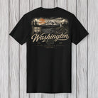 A black short-sleeve t-shirt with a detailed graphic print on the back, featuring a mountain landscape, a mountain sign, and the text 'Washington' in a cursive font, displayed against a light gray wood plank background.
