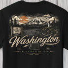 A black short-sleeve t-shirt featuring a detailed back graphic of Mount Hood, Oregon, with the text 'The Evergreen State' and 'Washington'. The design includes an orange sun, pine trees, a lighthouse, and a map outline of Washington, shown against a light gray wood plank background.
