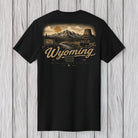 A black short-sleeve t-shirt with a detailed vintage-style Wyoming landscape graphic on the back, featuring a mountain scene, a sun, and the text 'Wyoming' in a cursive font, displayed against a light gray wood plank background.