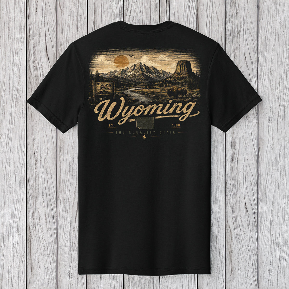 A black short-sleeve t-shirt with a detailed vintage-style Wyoming landscape graphic on the back, featuring a mountain scene, a sun, and the text 'Wyoming' in a cursive font, displayed against a light gray wood plank background.