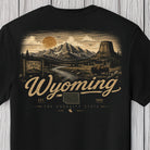 A black short-sleeve t-shirt with a detailed Wyoming state graphic on the front, featuring a mountain landscape, a bison, and the text 'Wyoming' in a vintage serif font, displayed against a light gray wood plank background.