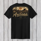 A black short-sleeve t-shirt with a detailed Arizona desert landscape graphic on the front, featuring the text 'Arizona' and 'The Grand Canyon State' in a vintage style, displayed flat against a light gray wood plank background.