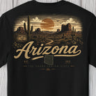 A black short-sleeve t-shirt featuring a detailed desert landscape graphic on the front. The design depicts Grand Canyon rock formations, saguaro cacti, a winding path, and a large orange sun in a cloudy sky, with 'Arizona' written in a textured beige script across the center.