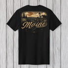 A black short-sleeve t-shirt with a vintage-style Maine graphic on the back, featuring a lighthouse, sunset, and the word 'Maine' in large script, displayed against a white wood plank background.