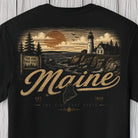A black short-sleeve t-shirt featuring a vintage-style Maine graphic print on the front. The design includes a lighthouse, pine trees, a sunset, and the words 'Maine', 'The Pine Tree State', and 'Est.