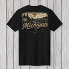 A black short-sleeve t-shirt with a vintage-style back graphic featuring a Michigan landscape, a large moon, and the text 'Michigan' and 'The Great Lakes Coast'. The shirt is displayed flat against a light gray wood plank background.