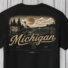 A black short-sleeve t-shirt with a vintage-style Michigan graphic print on the front, featuring a lighthouse, lake, pine trees, and the word 'Michigan' in large textured lettering, shown against a light wood plank background.