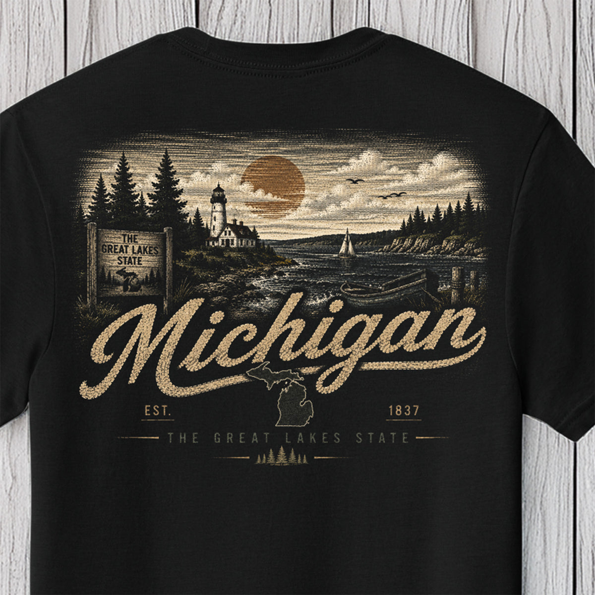 A black short-sleeve t-shirt with a vintage-style Michigan graphic print on the front, featuring a lighthouse, lake, pine trees, and the word 'Michigan' in large textured lettering, shown against a light wood plank background.