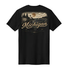 A black short-sleeve t-shirt shown from the back, featuring a vintage-style scenic graphic of Michigan's Great Lakes Coast with a lighthouse, sunset, and large 'Michigan' text in tan.