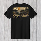 A black short-sleeve t-shirt with a vintage-style Minnesota landscape graphic on the back, featuring a sunset, trees, and the word 'Minnesota' in a cursive font, displayed against a light gray wood plank background.