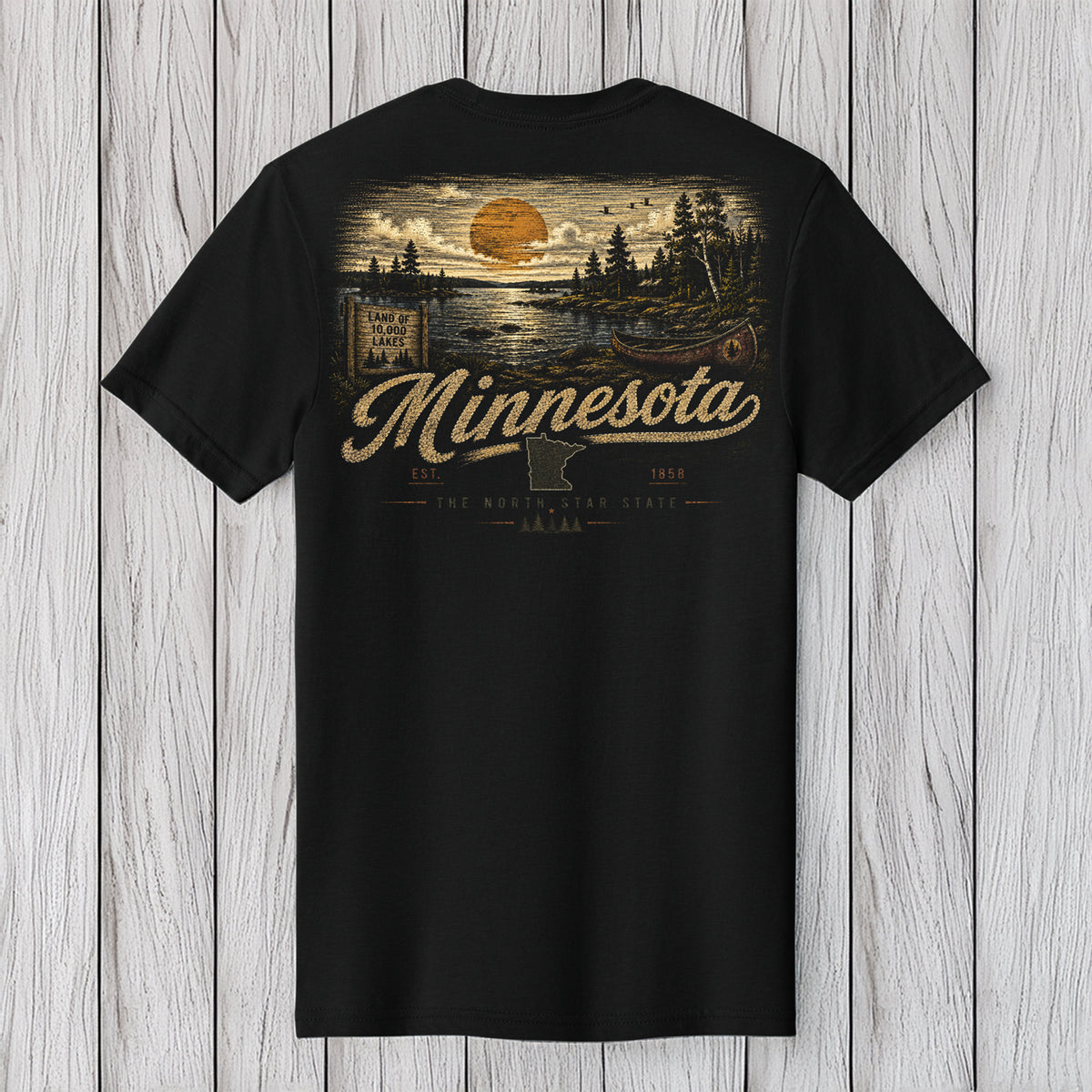 A black short-sleeve t-shirt with a vintage-style Minnesota landscape graphic on the back, featuring a sunset, trees, and the word 'Minnesota' in a cursive font, displayed against a light gray wood plank background.