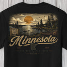 A black short-sleeve t-shirt with a large vintage-style Minnesota graphic on the back, featuring a sunset over a lake, trees, a canoe, and the word 'Minnesota' in textured lettering, shown against a light wood plank background.