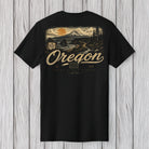 A black short-sleeve t-shirt with a large vintage-style Oregon graphic on the front, featuring a mountain, sunset, and the word 'Oregon' in beige, displayed against a light gray wood plank background.