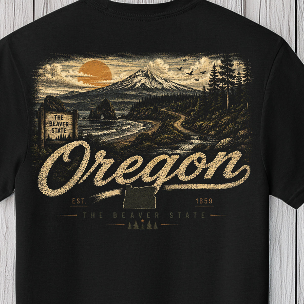 A black short-sleeve t-shirt with a large vintage-style Oregon graphic on the front, featuring a mountain landscape, river, and the word 'Oregon' in large textured lettering, shown against a light wood plank background.