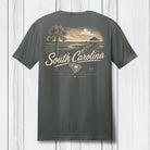 A dark gray t-shirt shown from the back, featuring a vintage-style South Carolina graphic with a palm tree, beach scene, and text, displayed against a white wood plank background.