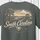 A dark gray t-shirt featuring a vintage-style South Carolina graphic print on the front, showing a palm tree, a boardwalk pier, and the text 'South Carolina' and 'The Palmetto State'.