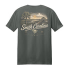 A dark gray t-shirt shown from the back, featuring a vintage-style graphic print of a South Carolina beach scene with a palm tree, sunset, and the text 'South Carolina' in cream lettering.