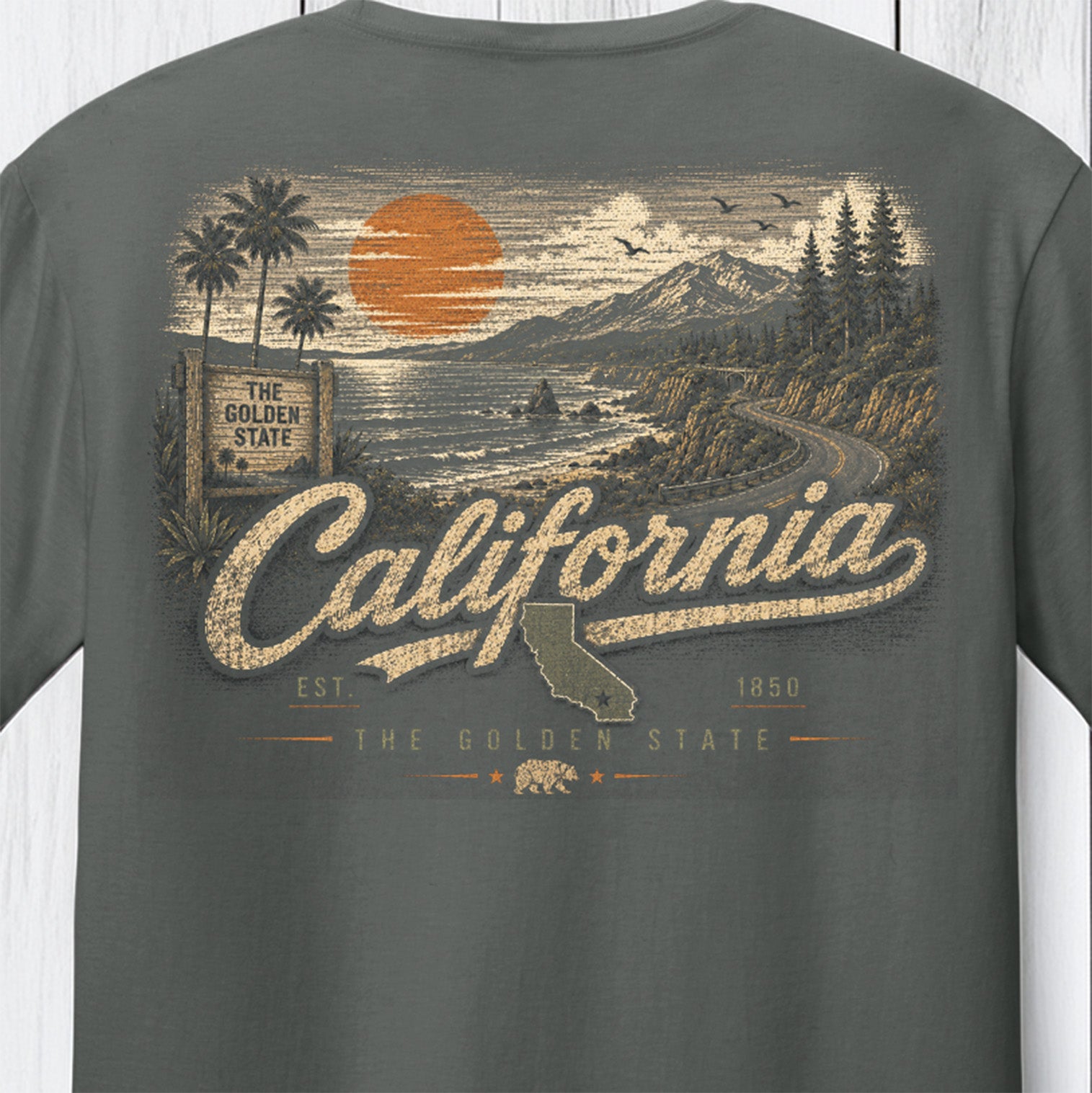 A dark gray t-shirt featuring a large vintage-style California graphic print on the front. The design includes a sunset over the ocean, mountains, palm trees, a winding road, and the words 'California' and 'The Golden State' in distressed cream and orange tones.
