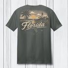 A dark gray t-shirt shown from the back, featuring a vintage-style Florida graphic print in beige and yellow tones with palm trees, a lighthouse, and the word 'Florida' in script, set against a white wood plank background.
