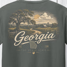 A close-up of a dark gray t-shirt showing a detailed vintage-style graphic print on the back. The graphic features a scenic Georgia landscape with a Spanish mossy oak tree, a dirt road, a sunset, and the word 'Georgia' in large script over a map of Georgia.