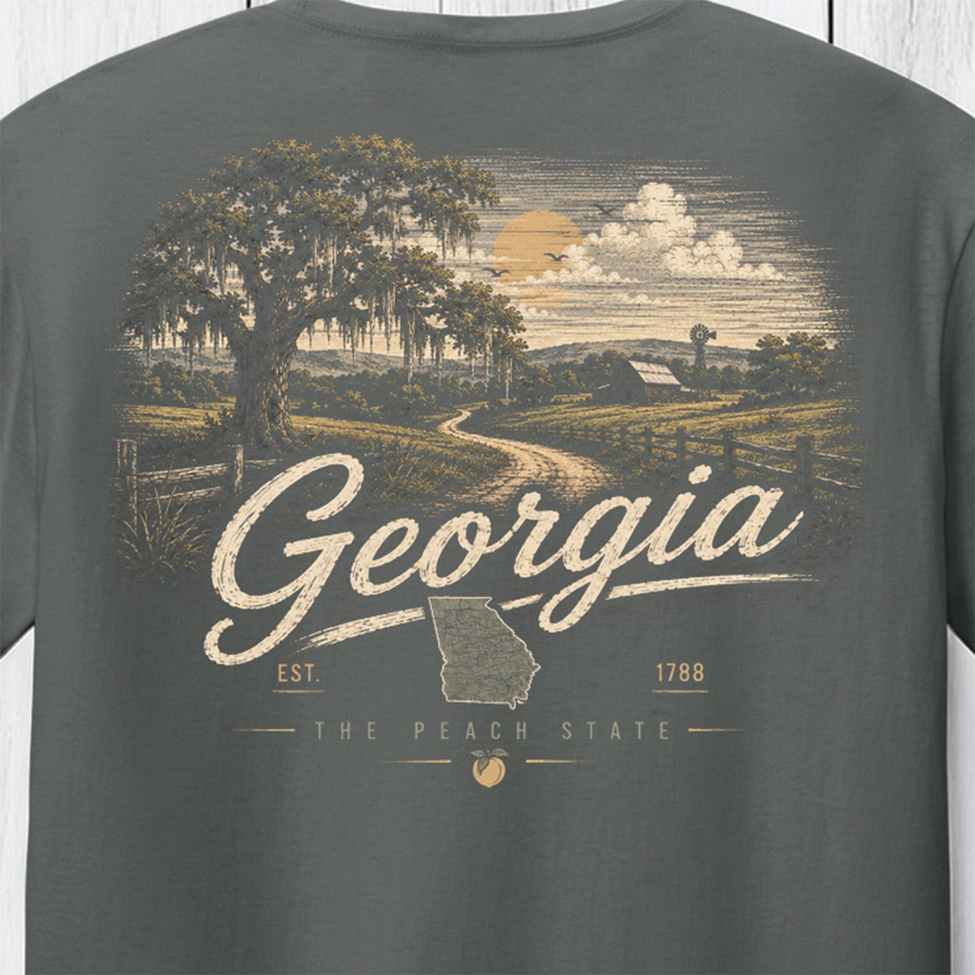 A close-up of a dark gray t-shirt showing a detailed vintage-style graphic print on the back. The graphic features a scenic Georgia landscape with a Spanish mossy oak tree, a dirt road, a sunset, and the word 'Georgia' in large script over a map of Georgia.