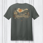 A dark gray t-shirt shown from the back, featuring a vintage-style Hawaii graphic with a sunset, mountains, palm trees, and the word 'Hawaii' in cream script, displayed against a white wood plank background.