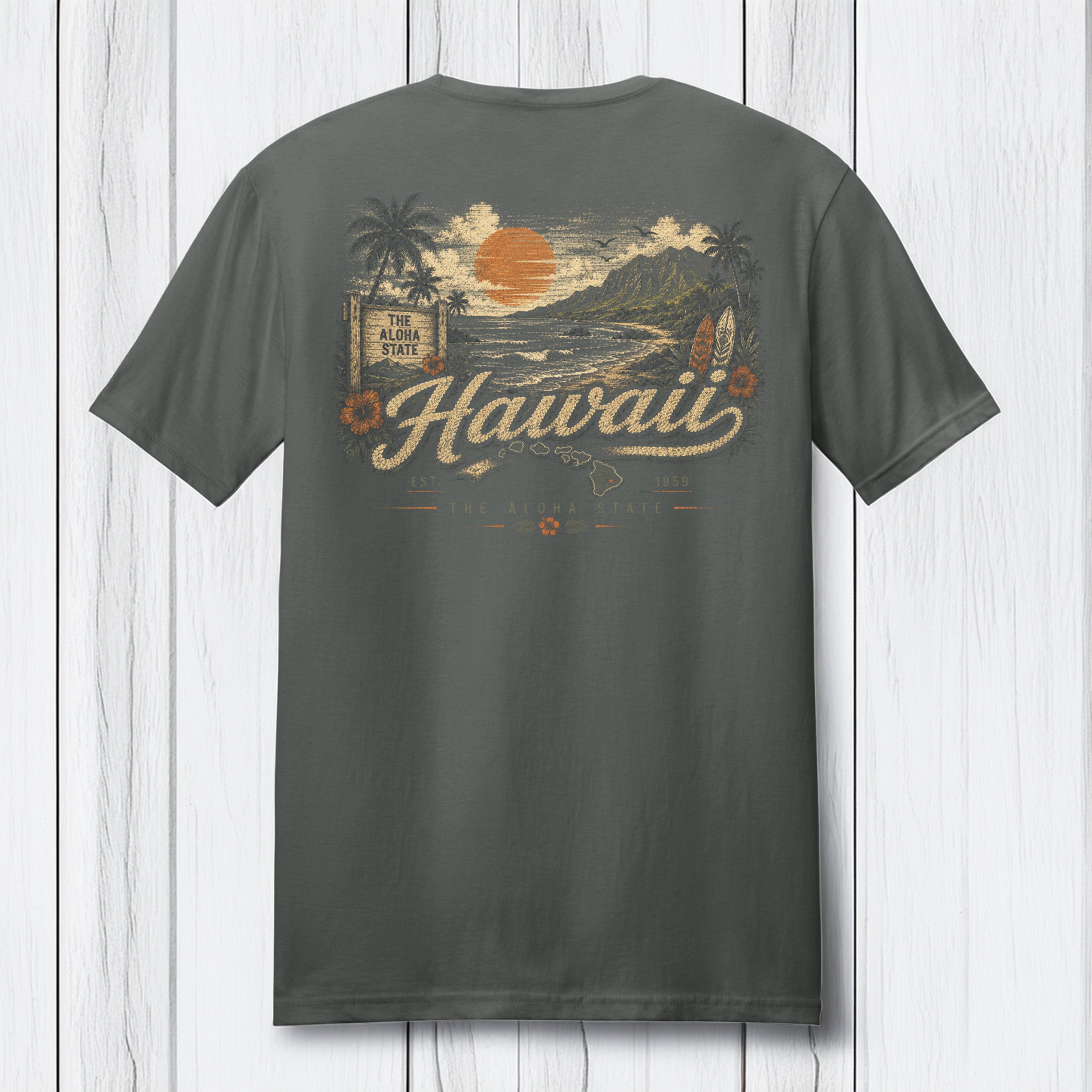 A dark gray t-shirt shown from the back, featuring a vintage-style Hawaii graphic with a sunset, mountains, palm trees, and the word 'Hawaii' in cream script, displayed against a white wood plank background.
