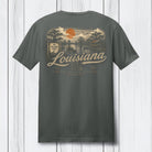 A dark gray t-shirt shown from the back, featuring a vintage-style Louisiana graphic with an orange sun, trees, and the word 'Louisiana' in beige script, displayed against a white wood plank background.