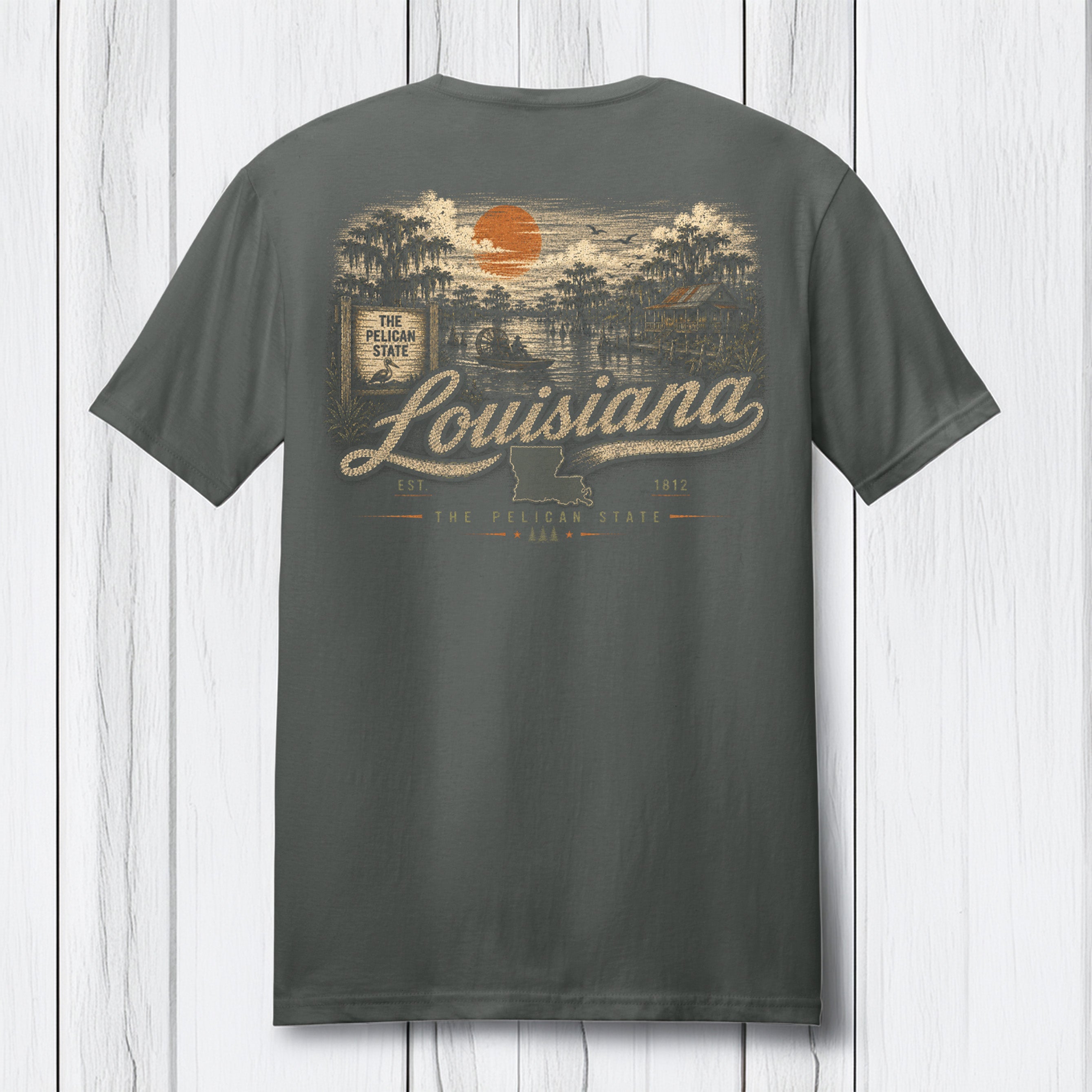 A dark gray t-shirt shown from the back, featuring a vintage-style Louisiana graphic with an orange sun, trees, and the word 'Louisiana' in beige script, displayed against a white wood plank background.