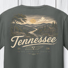 A dark gray t-shirt featuring a vintage-style Tennessee landscape graphic on the front, with a watercolor-style illustration of a mountain valley, a waterwheel, and the word 'Tennessee' in large script.
