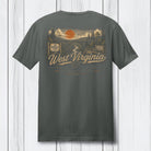 A dark gray t-shirt shown from the back, featuring a vintage-style West Virginia graphic with an orange sun, river, bridge, and 'West Virginia' text, displayed against a white wood plank background.