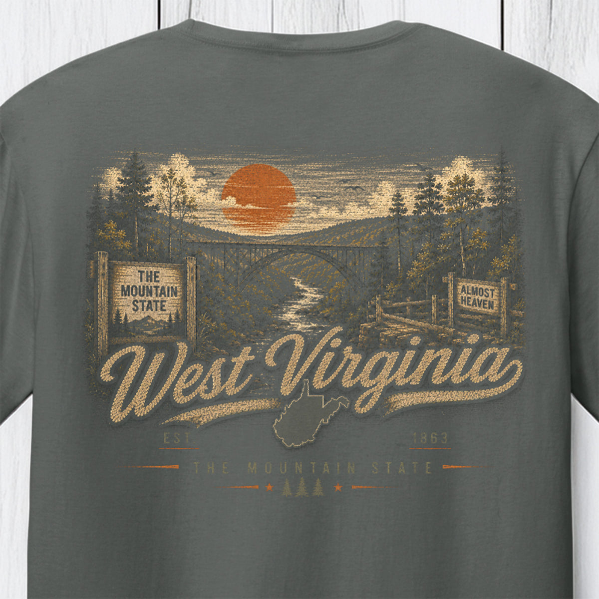 A dark gray t-shirt featuring a vintage-style West Virginia graphic print with an orange sun, bridge, trees, and the words 'West Virginia' and 'The Mountain State'.
