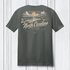 A dark gray t-shirt shown from the back, featuring a vintage-style North Carolina graphic with a sunset, lighthouse, waves, and the text 'North Carolina' in cream and gold, displayed against a white wood plank background.