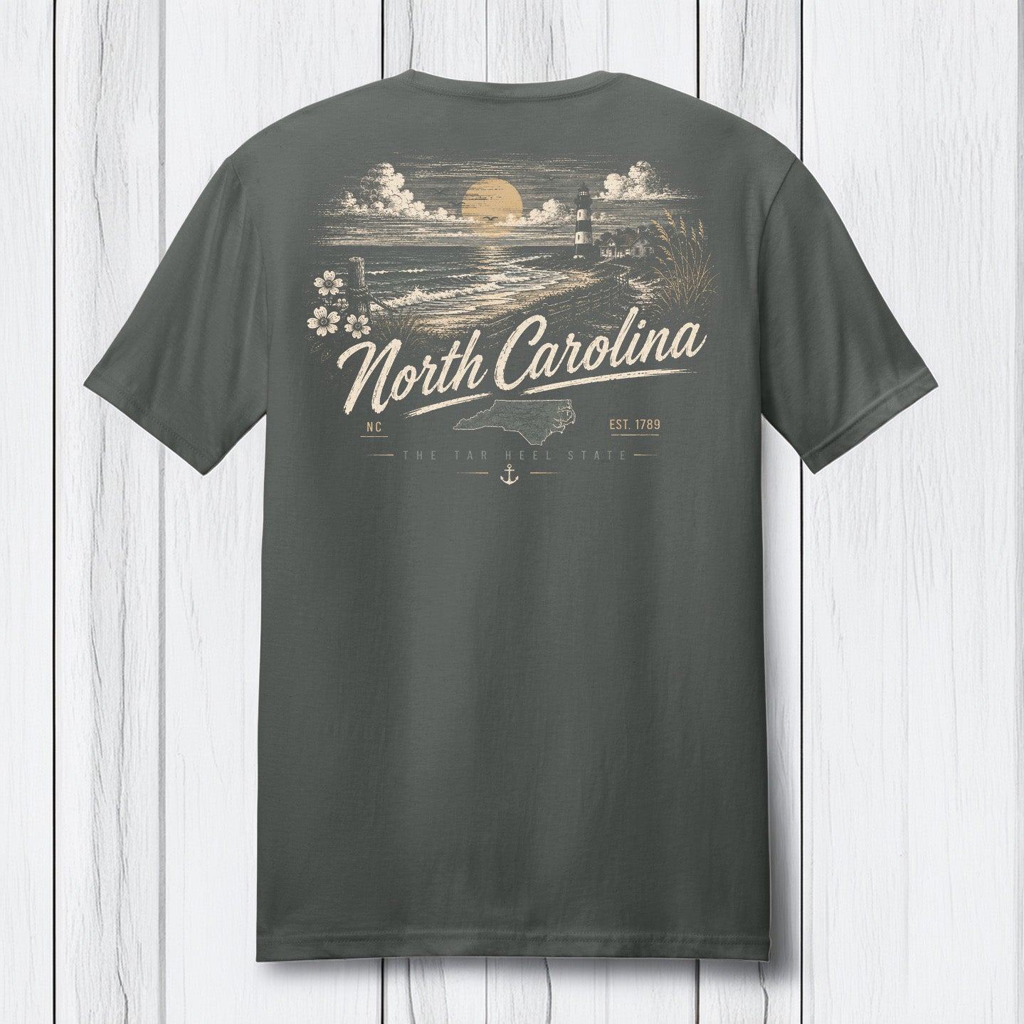 A dark gray t-shirt shown from the back, featuring a vintage-style North Carolina graphic with a sunset, lighthouse, waves, and the text 'North Carolina' in cream and gold, displayed against a white wood plank background.