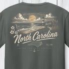 A dark gray t-shirt showing the back graphic print with a coastal sunset scene, lighthouse, and the text 'North Carolina' in a vintage script.