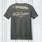 A dark gray t-shirt shown from the back, featuring a vintage-style North Carolina graphic with a mountain landscape, pine trees, a sun, and the text 'North Carolina' in cream lettering, displayed against a white wood plank background.