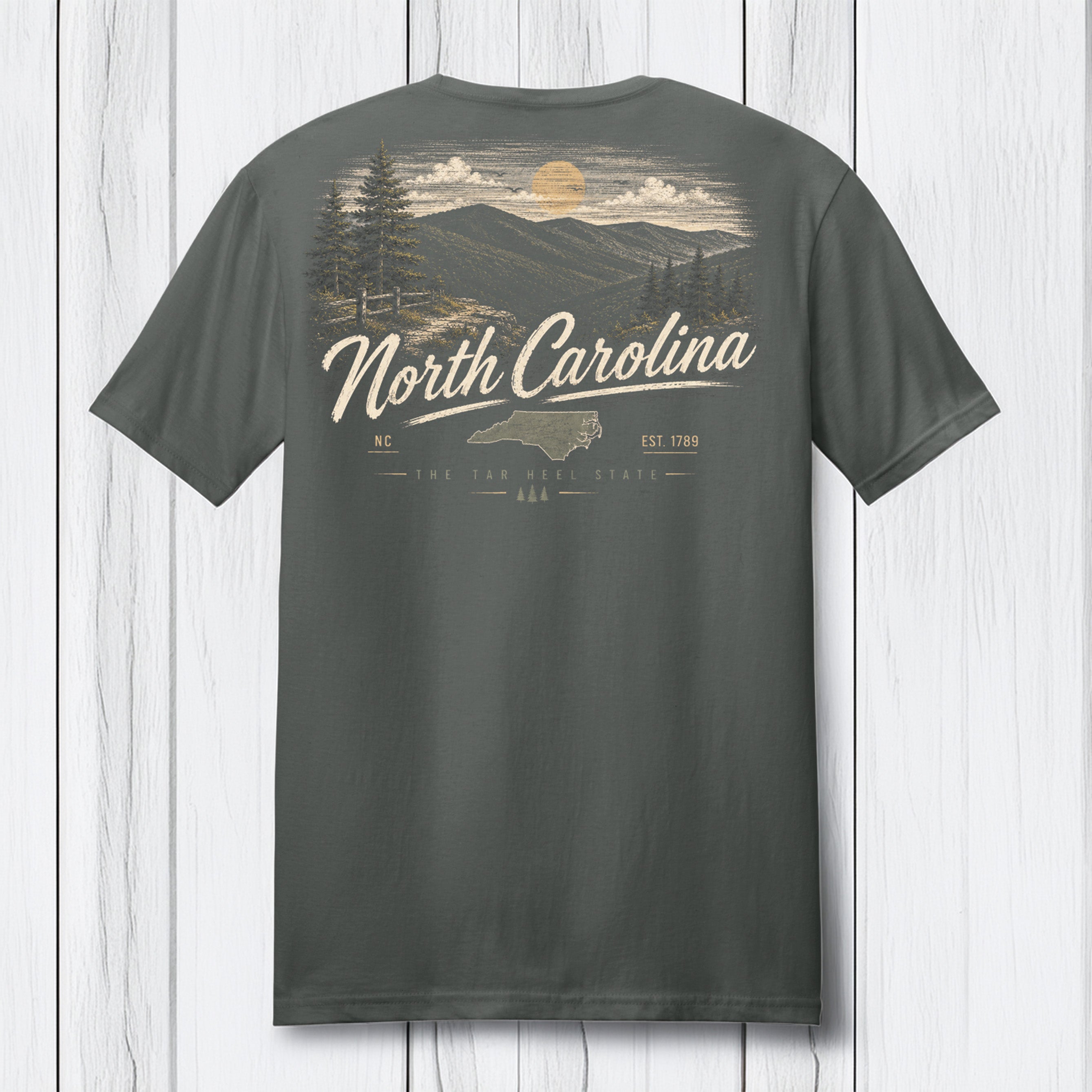 A dark gray t-shirt shown from the back, featuring a vintage-style North Carolina graphic with a mountain landscape, pine trees, a sun, and the text 'North Carolina' in cream lettering, displayed against a white wood plank background.