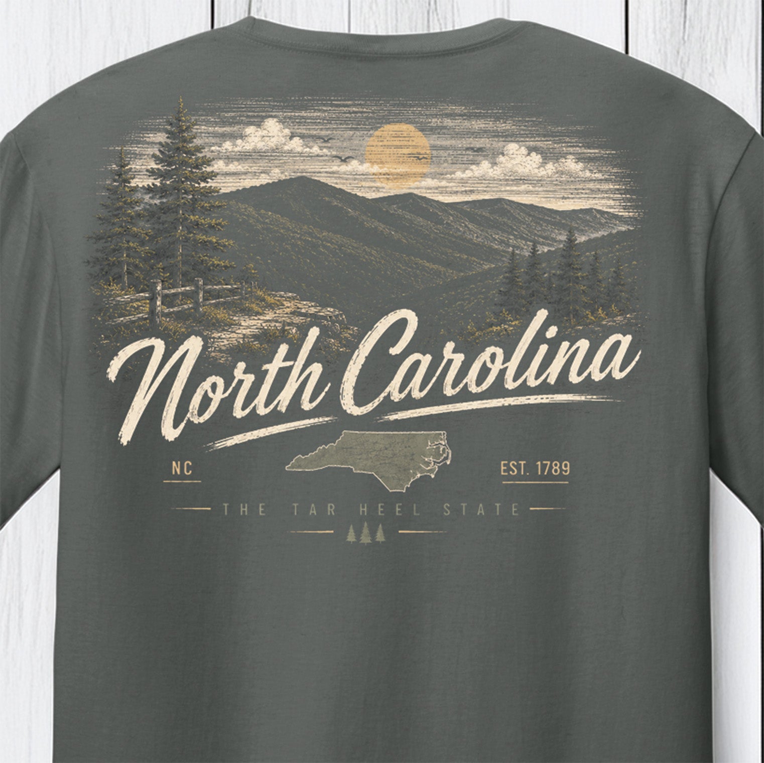 A dark gray t-shirt showing the back graphic print with a vintage-style North Carolina landscape, featuring mountains, pine trees, a sun, and the text 'North Carolina' and 'The Tar Heel State'.