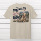 Sand-colored unisex t-shirt featuring "The Mountains Are Better With Dogs" graphic with a happy Golden Retriever sitting in the Blue Ridge Mountains, displayed on a rustic wood background.