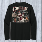 A black long-sleeve t-shirt shown from the back, featuring a large Christmas-themed graphic print with a snowman, campfire, lanterns, and the text 'Chillin' Through the Holidays' and 'Black Frog Apparel'. The shirt is displayed flat against a light gray wood-plank background.
