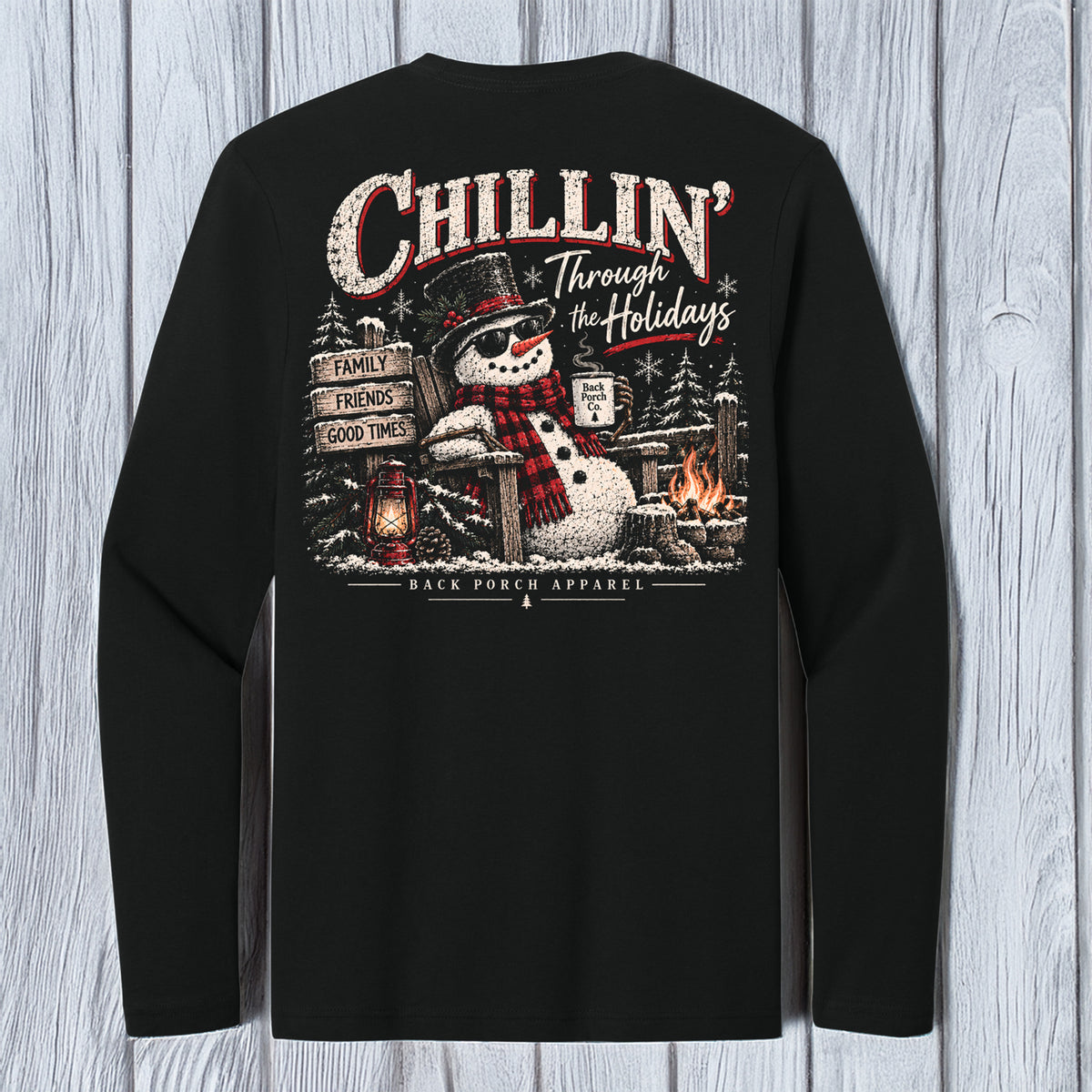A black long-sleeve t-shirt shown from the back, featuring a large Christmas-themed graphic print with a snowman, campfire, lanterns, and the text 'Chillin' Through the Holidays' and 'Black Frog Apparel'. The shirt is displayed flat against a light gray wood-plank background.