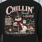 A close-up of a black sweatshirt featuring a large festive graphic on the front. The design shows a snowman wearing a top hat and red plaid scarf, sitting by a campfire with a mug that reads 'Back Porch Co.